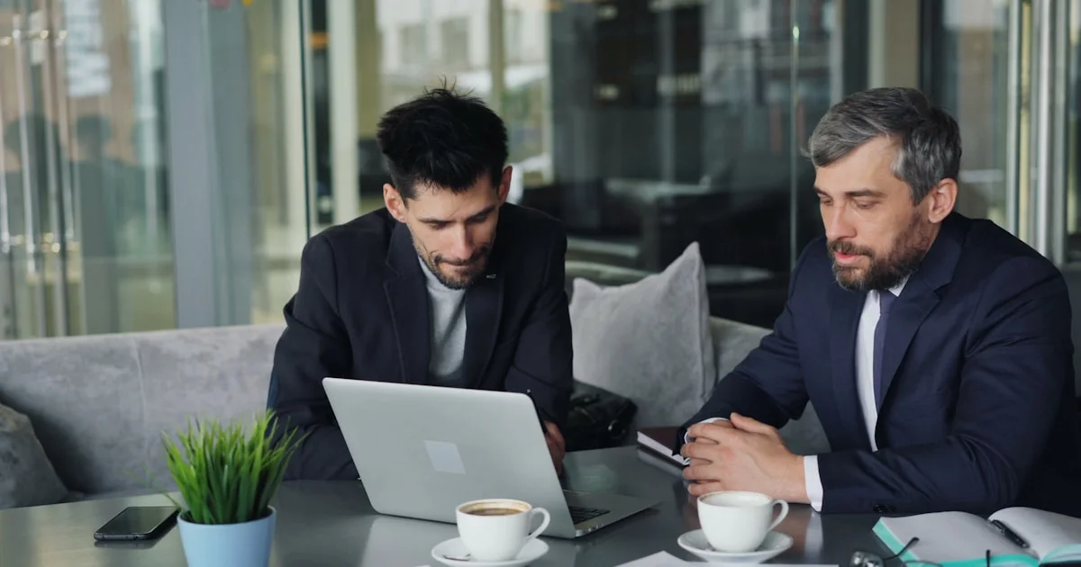 two men sitting at a table looking at a laptop