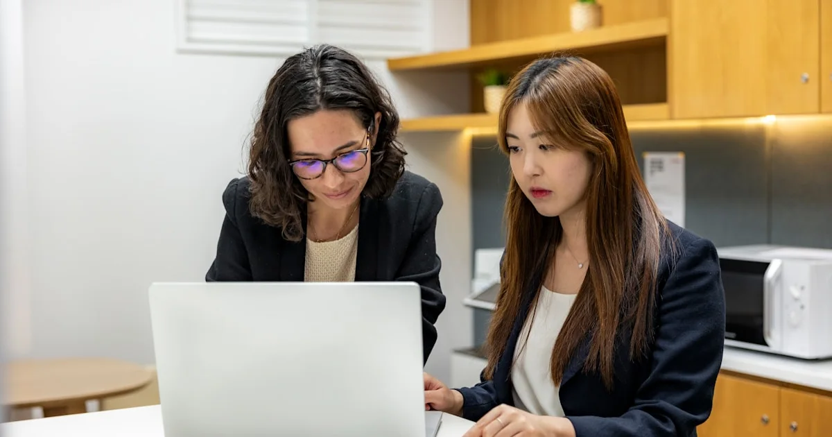 two women sitting at a table looking at a laptop
