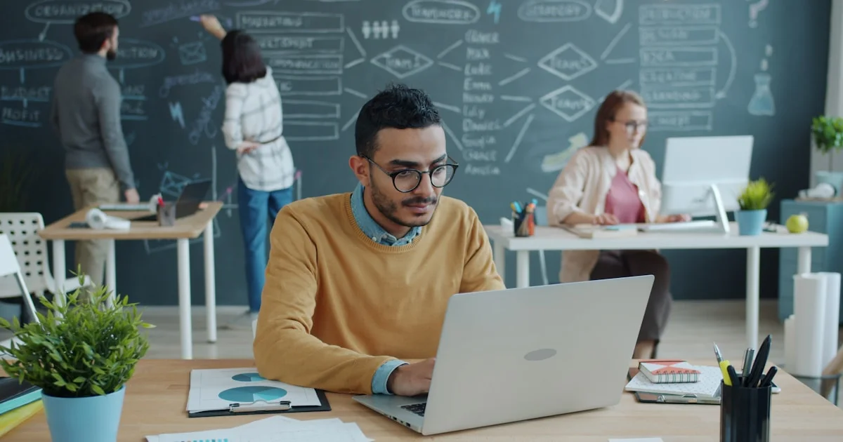 Man working on laptop in modern office with colleagues.